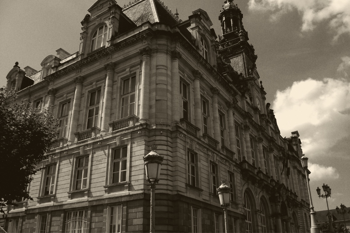 The Hôtel de Ville of Limoges, a tall nineteenth-century stone town hall with a slate mansard roof and a belfry, seen from a corner below with old street lamps in front and clouds behind, in a dark ink and bone duotone