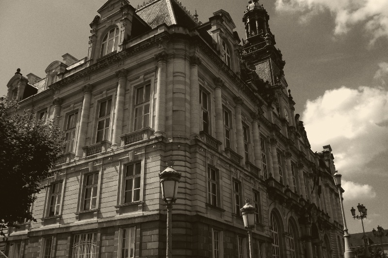 The Hôtel de Ville of Limoges, a tall nineteenth-century stone town hall with a slate mansard roof and a belfry, seen from a corner below with old street lamps in front and clouds behind, in a dark ink and bone duotone