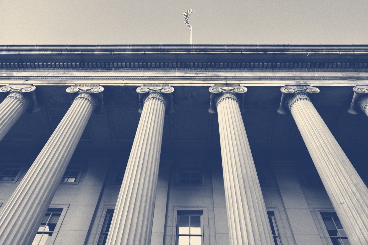 The colonnade of the United States Treasury building seen from below with a flag on the roofline, in a slate and bone duotone