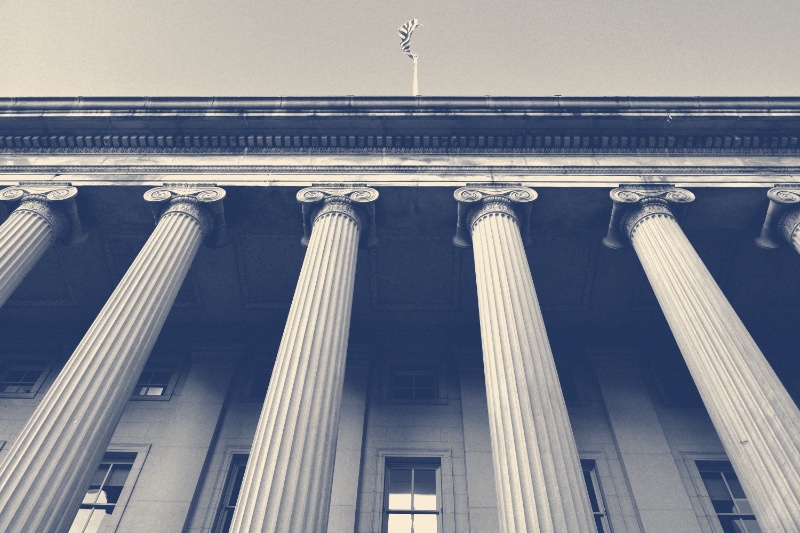 The colonnade of the United States Treasury building seen from below with a flag on the roofline, in a slate and bone duotone
