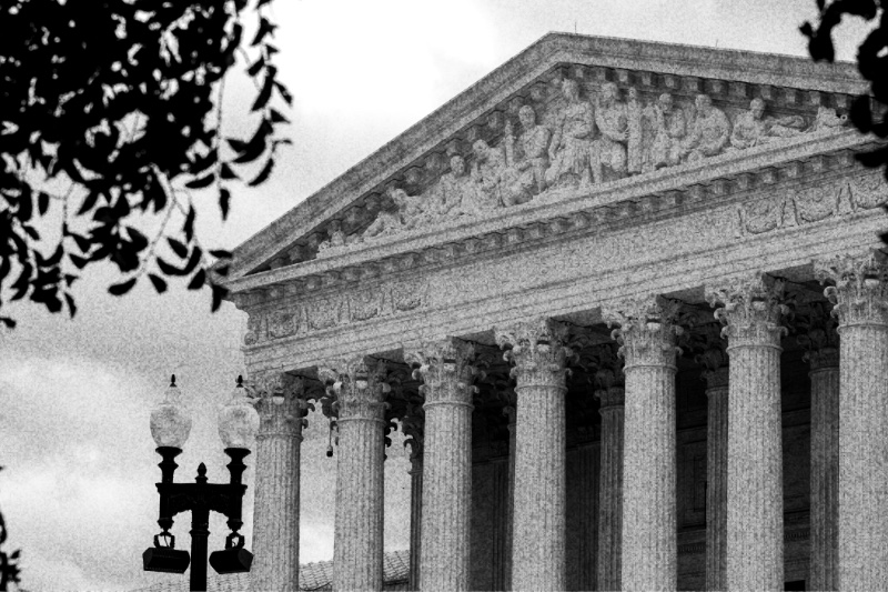 The pediment and columns of the United States Supreme Court under a heavy sky, seen past a street lamp, in high contrast black and white with coarse grain