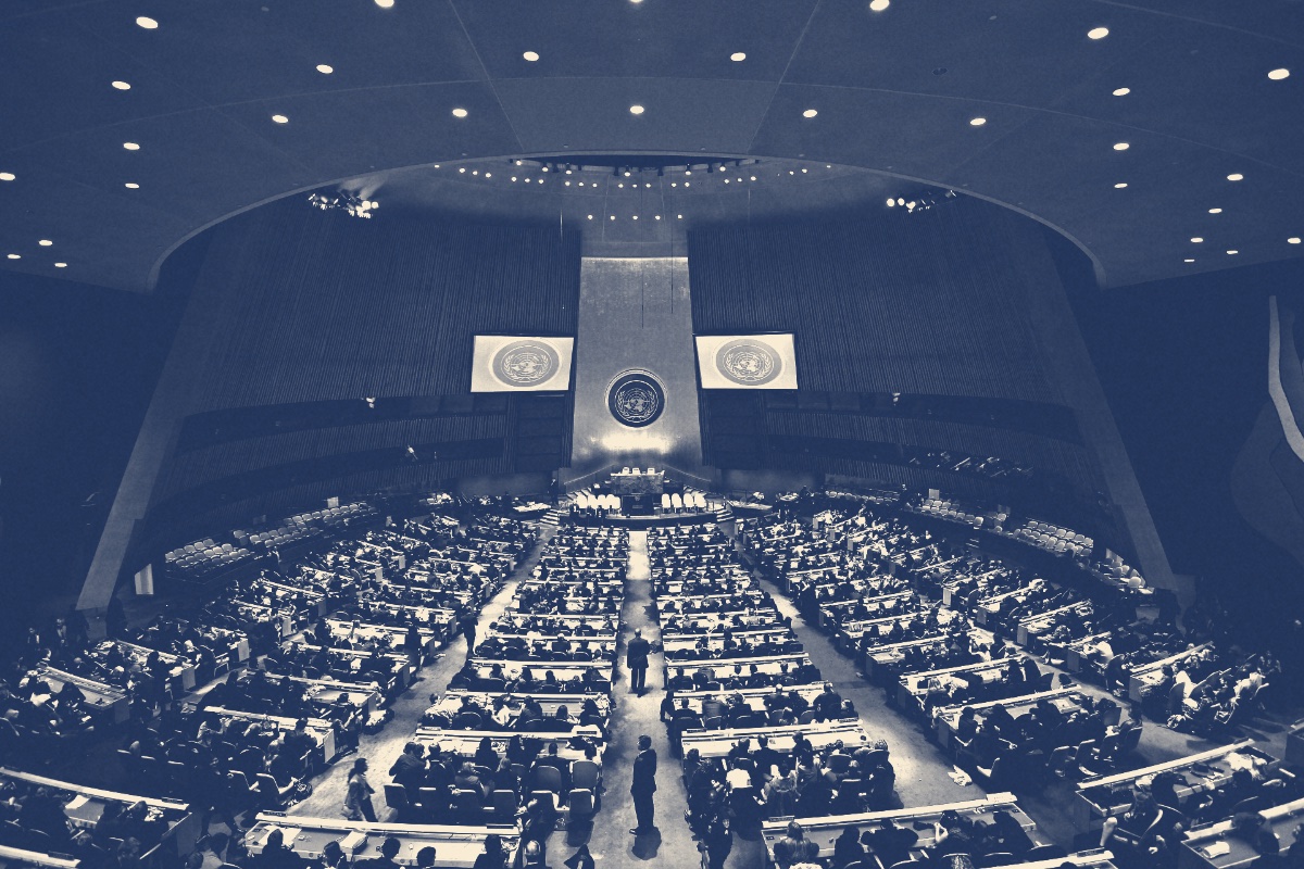 The United Nations General Assembly hall in New York seen from the rear of the chamber, rows of desks facing the marble rostrum, in a slate and bone duotone