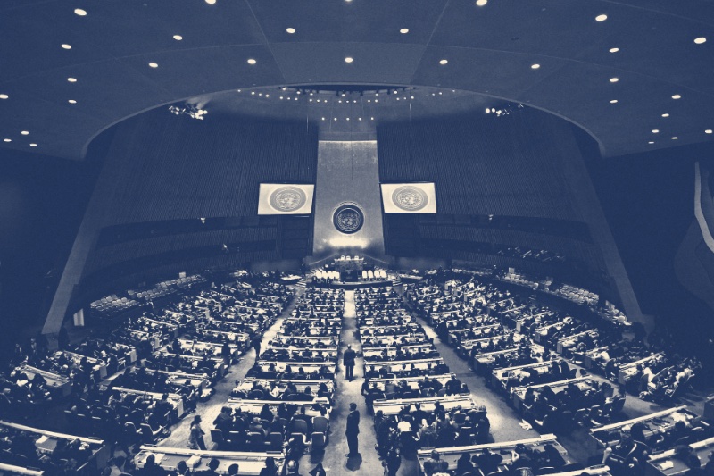 The United Nations General Assembly hall in New York seen from the rear of the chamber, rows of desks facing the marble rostrum, in a slate and bone duotone