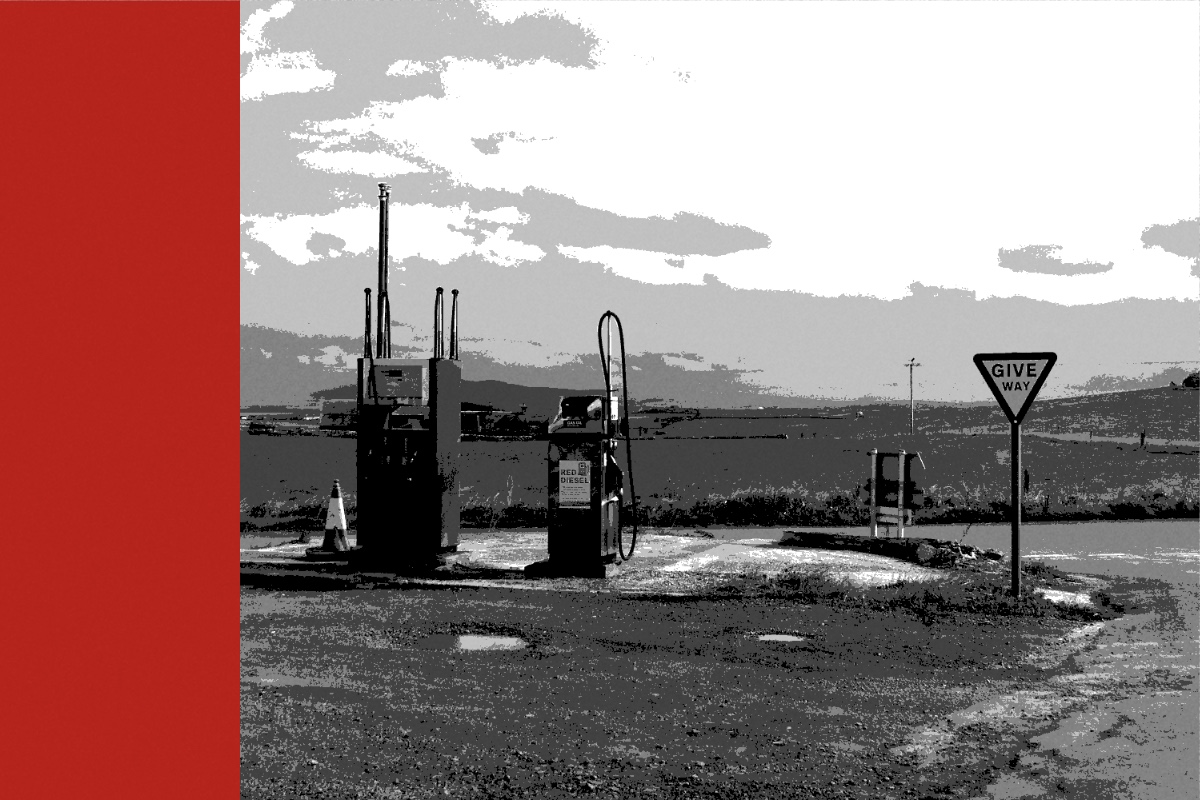 A red diesel pump and a second fuel pump on a gravel forecourt beside open fields, a give way sign at the right, in near black and white with a flat red bar down the left edge of the frame