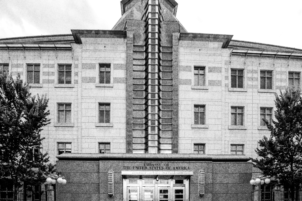 The entrance of the United States Embassy in Ottawa, a stone facade with a tall glass and metal central tower over a granite base marked Embassy of the United States of America, trees at either side, in grainy high-contrast black and white