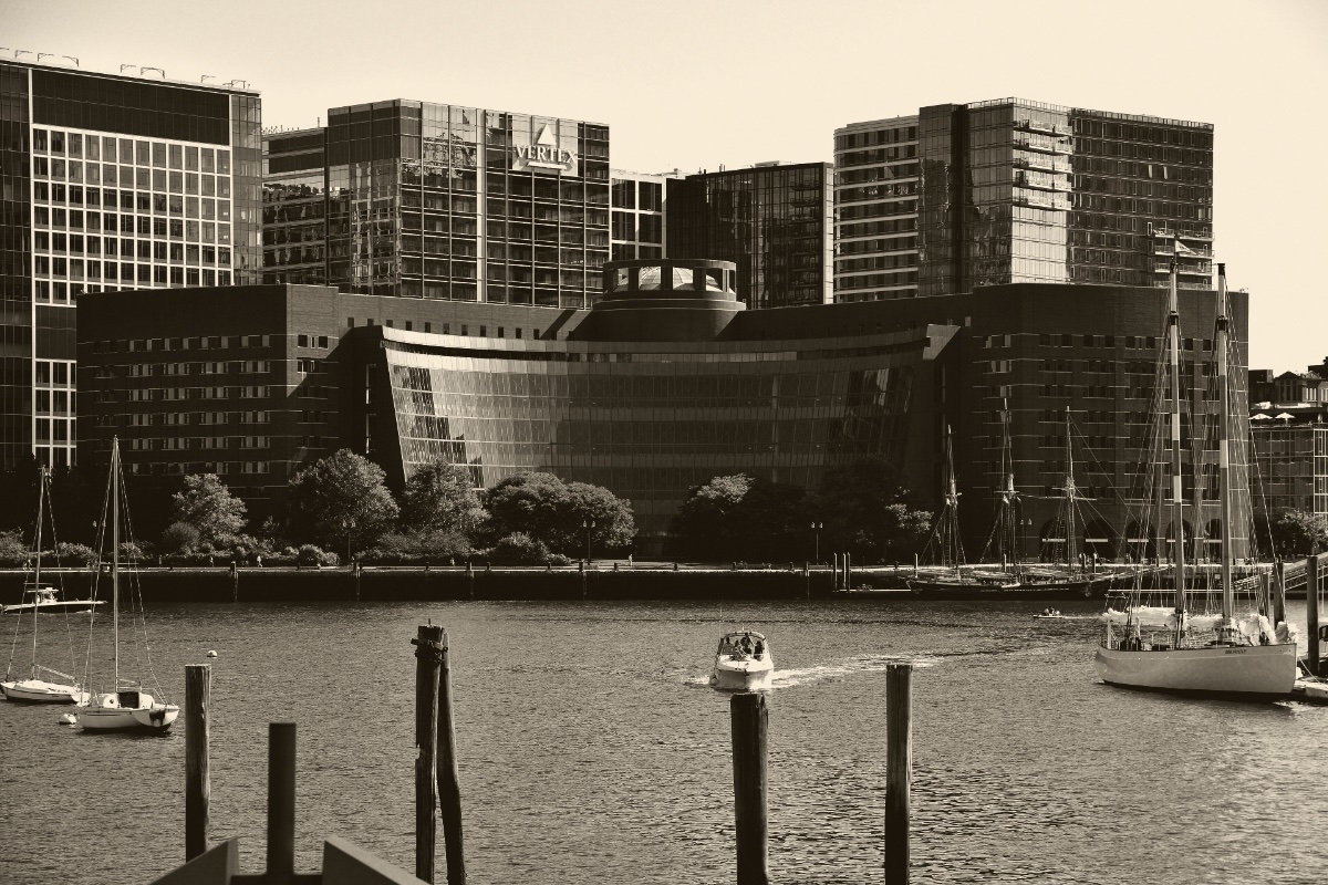 The John Joseph Moakley United States Courthouse in Boston seen across the harbour, a curved glass facade between brick wings with sailing boats moored in front, in an ink and bone duotone