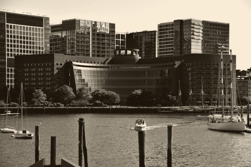 The John Joseph Moakley United States Courthouse in Boston seen across the harbour, a curved glass facade between brick wings with sailing boats moored in front, in an ink and bone duotone