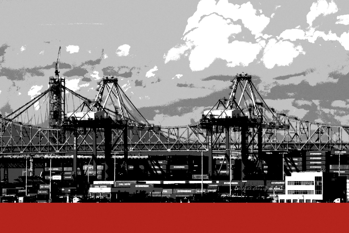 Container cranes over stacked containers at the Port of Oakland, a bridge behind them under heavy cloud, in near black and white with a flat red band along the foot of the frame