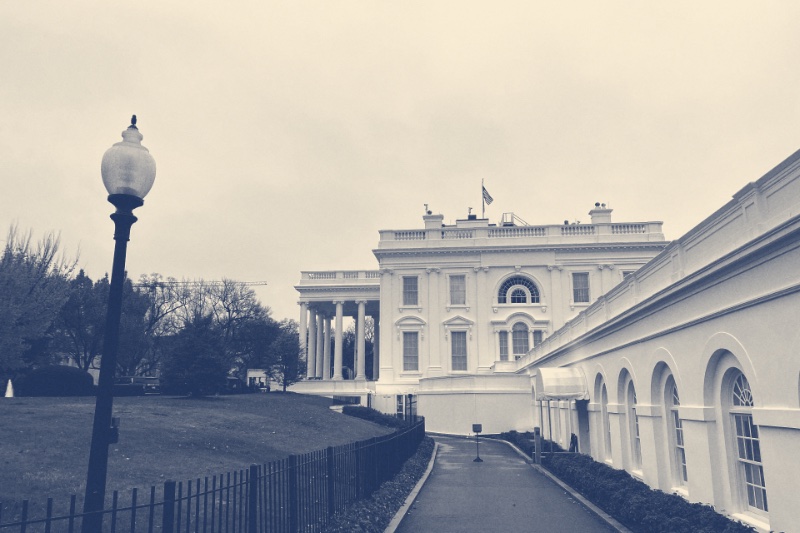 The path along the colonnade of the West Wing toward the White House, with a lamp post and iron fence on the left and an overcast sky, in a slate and bone duotone