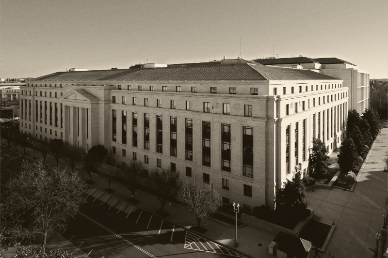 The Dirksen Senate Office Building in Washington seen from above, a long pale stone building with a green copper roof beside a tree-lined street under a clear sky, in an ink and bone duotone