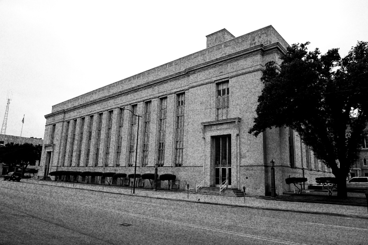 The United States Post Office and Courthouse in Wichita Falls, Texas, a long pale stone building with tall vertical windows behind a row of pilasters, seen from across an empty street with a tree at the corner, in grainy high-contrast black and white