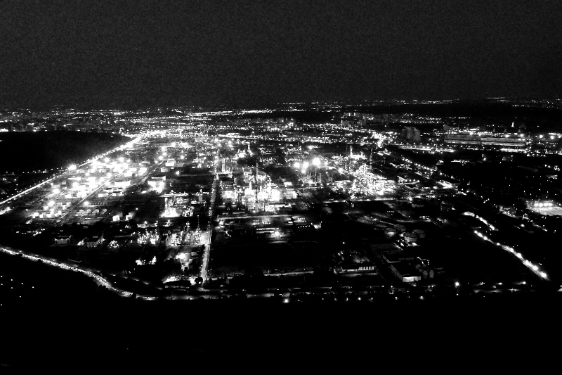 A Russian oil refinery at night seen from high above, a wide grid of bright lights and roads spreading across dark ground under a deep blue sky, in high contrast black and white with coarse grain