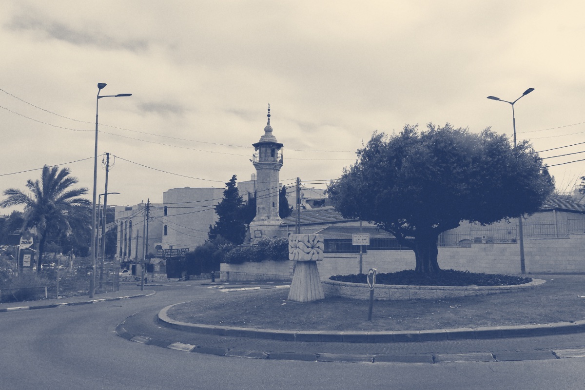 A roundabout in the Sheikh Jarrah neighbourhood of East Jerusalem, an olive tree and flower bed in the centre and the minaret of the Sheikh Jarrah mosque behind, in a slate and bone duotone