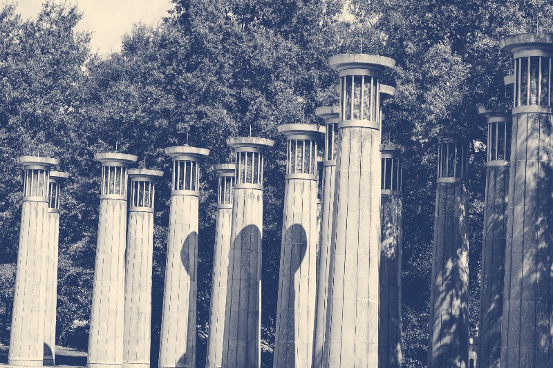The columns of the carillon at Bicentennial Capitol Mall in Nashville, each topped by an open bell housing, against a wall of trees, in a slate and bone duotone