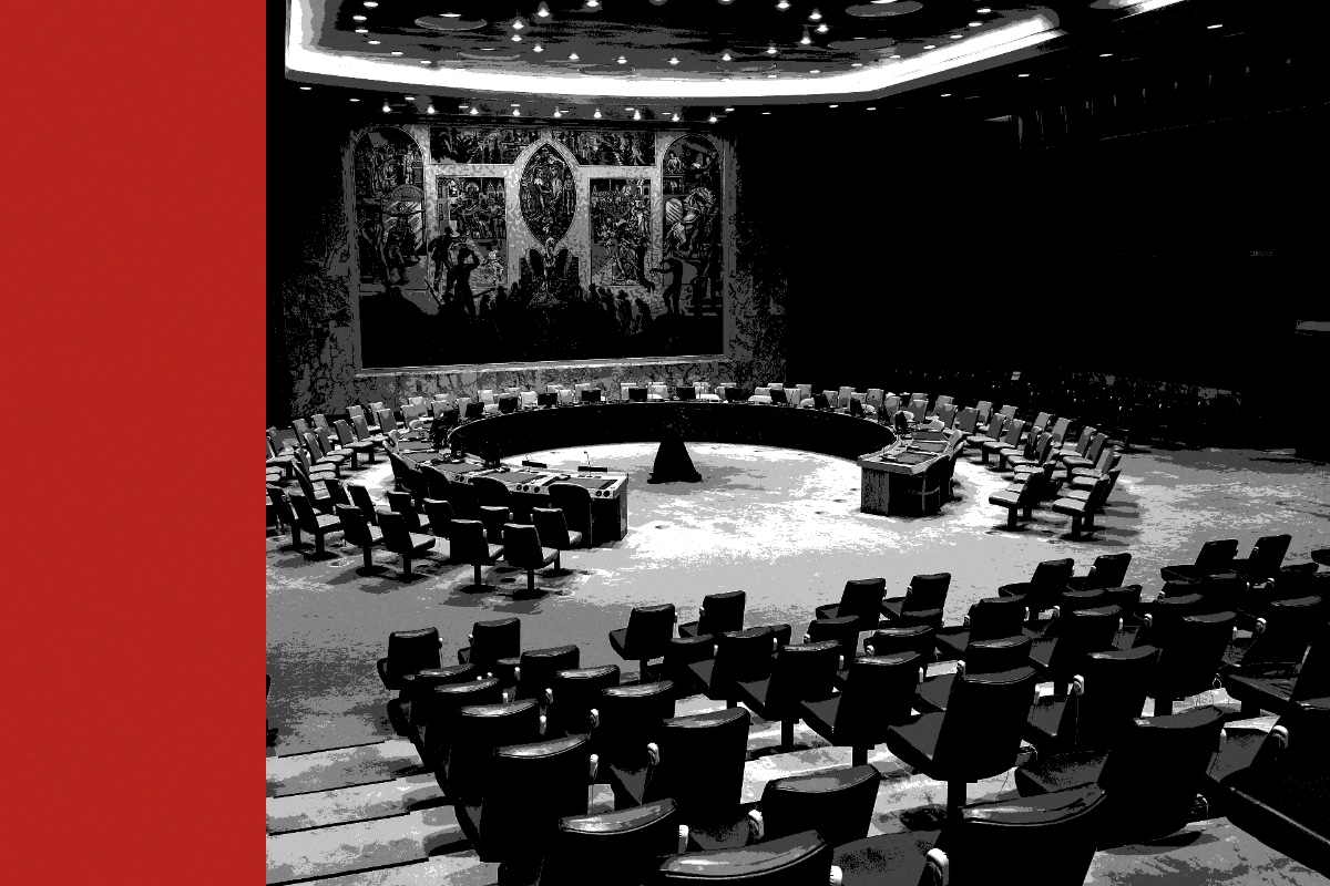 The horseshoe table of the United Nations Security Council chamber beneath its mural, rows of empty seats in the foreground, in near black and white with a flat red bar down the left edge of the frame