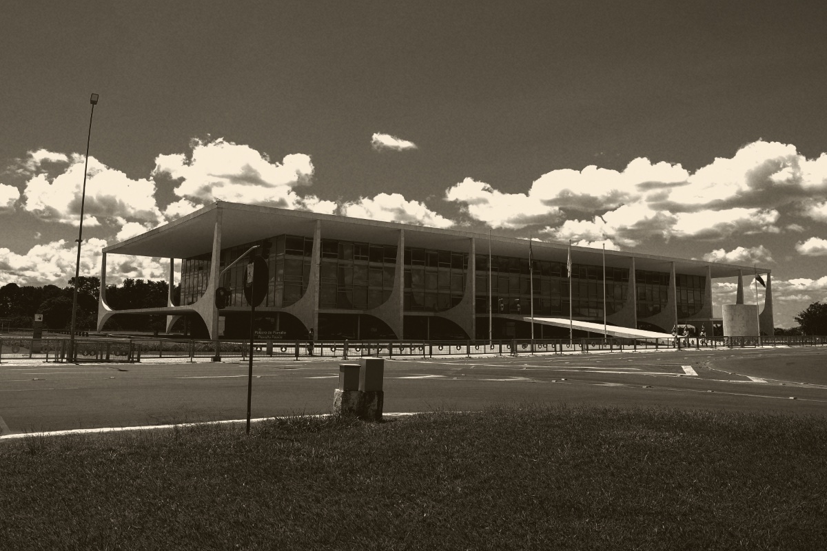 The Planalto Palace in Brasilia seen across an empty road and lawn, a long low glass building under a flat white roof on curved columns beneath a cloudy sky, in an ink and bone duotone