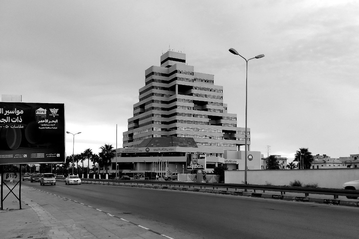 A stepped, terraced office block beside a wide road with traffic under an overcast sky, a billboard at the left edge of the frame, in black and white
