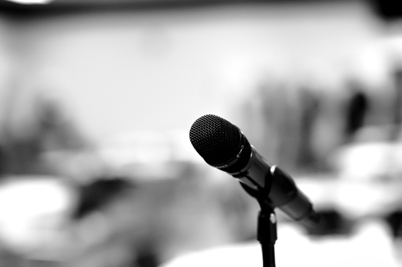 A single handheld microphone on a stand in sharp focus, with an empty room blurred behind it, in black and white