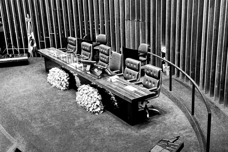 The presiding table of the Brazilian Federal Senate, a long wooden desk with empty grey leather chairs on a curved blue dais, flower arrangements at its front and tall vertical slats behind, in grainy high-contrast black and white