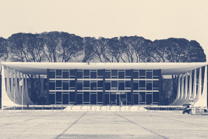 The Supreme Federal Court building in Brasília across an empty paved square, a low glass block under a flat roof held up by curved white columns, a line of trees behind, in a slate and bone duotone