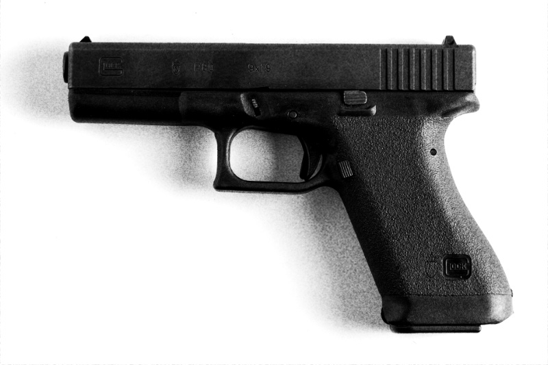 A Glock 17 pistol lying on its side against a plain pale background, its magazine seated in the grip, in grainy high-contrast black and white