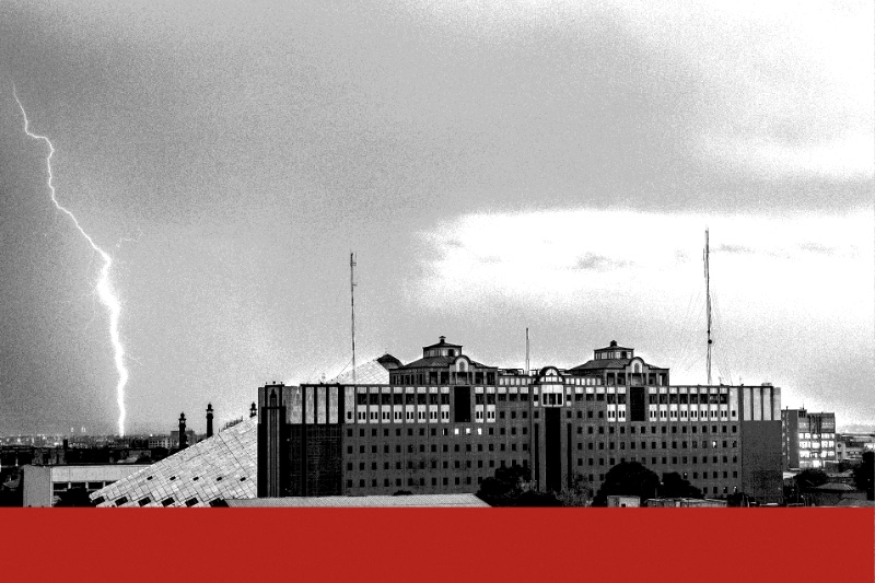 The Islamic Consultative Assembly building in Tehran, a long office block beside the sloping pyramid of its chamber, under storm clouds with a bolt of lightning at the left, in near black and white with a flat red band along the foot of the frame
