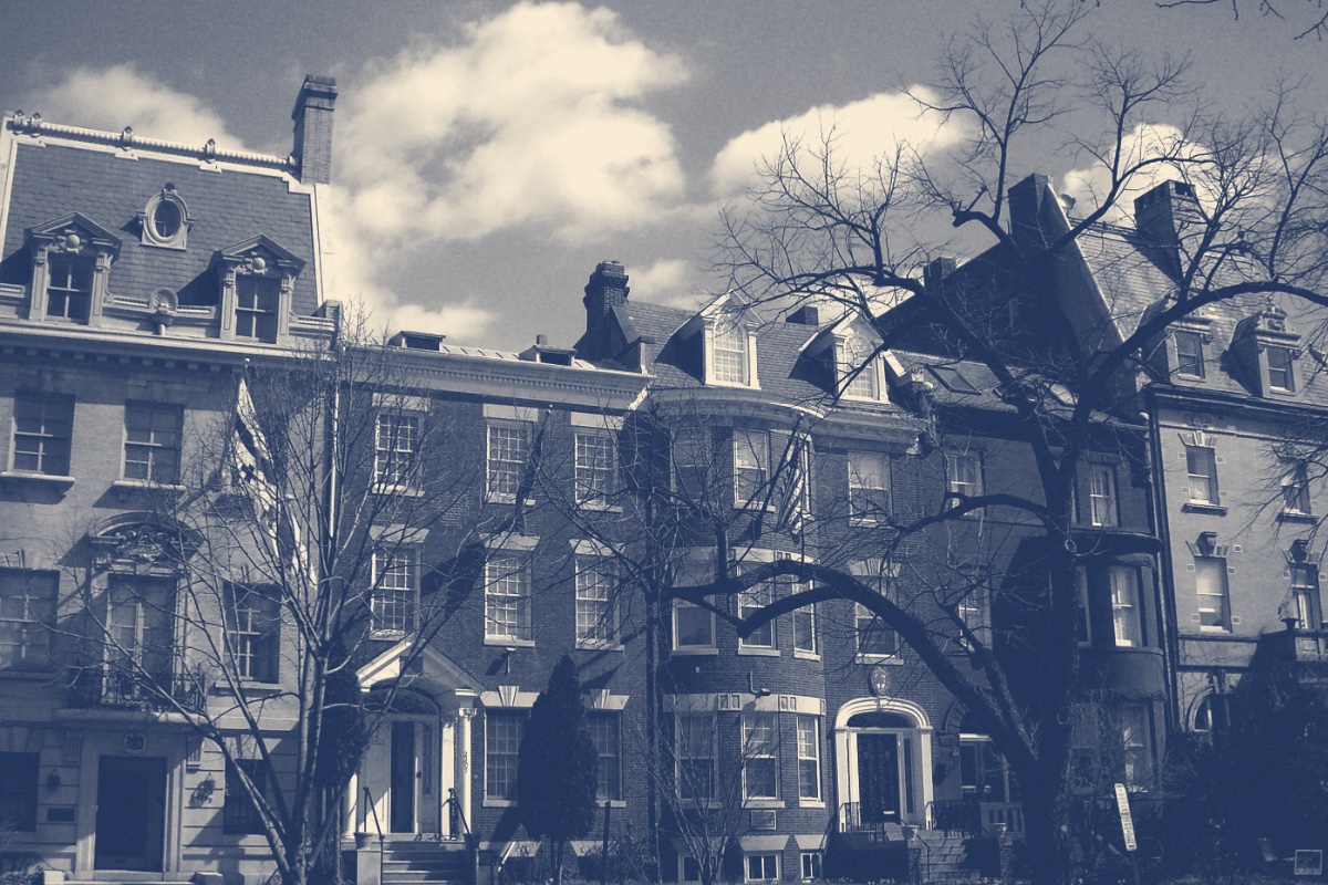 A row of brick and limestone townhouses on Massachusetts Avenue in Washington with flags hanging over two doorways and bare trees in front under a cloudy sky, in a slate and bone duotone