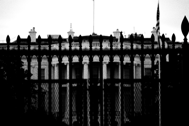 The White House seen through the spearheads and mesh of the north fence at dusk, in high contrast black and white with coarse grain