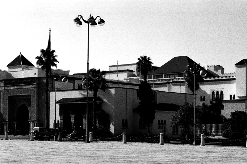 The Royal Palace in Rabat seen across a wide paved square, a long low pale building with green tiled roofs, a carved arched gateway and palm trees, a tall double street lamp in the foreground, in grainy high-contrast black and white