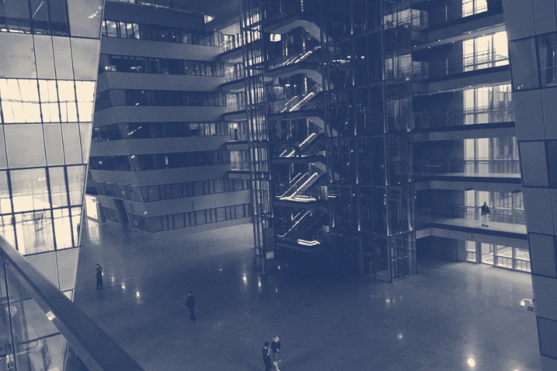 The glass atrium of NATO headquarters in Brussels with open staircases and a few figures crossing the floor, in a slate and bone duotone
