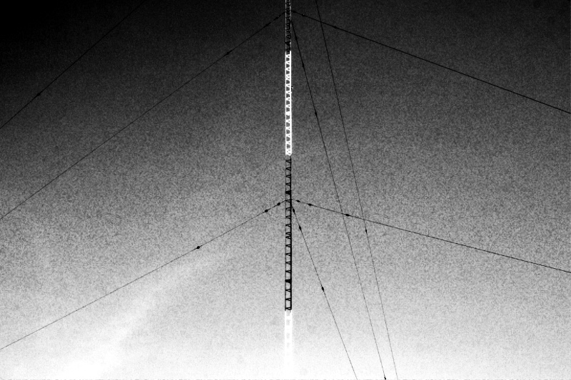 An AM radio mast radiator with guy wires against the sky, in high contrast black and white with coarse grain