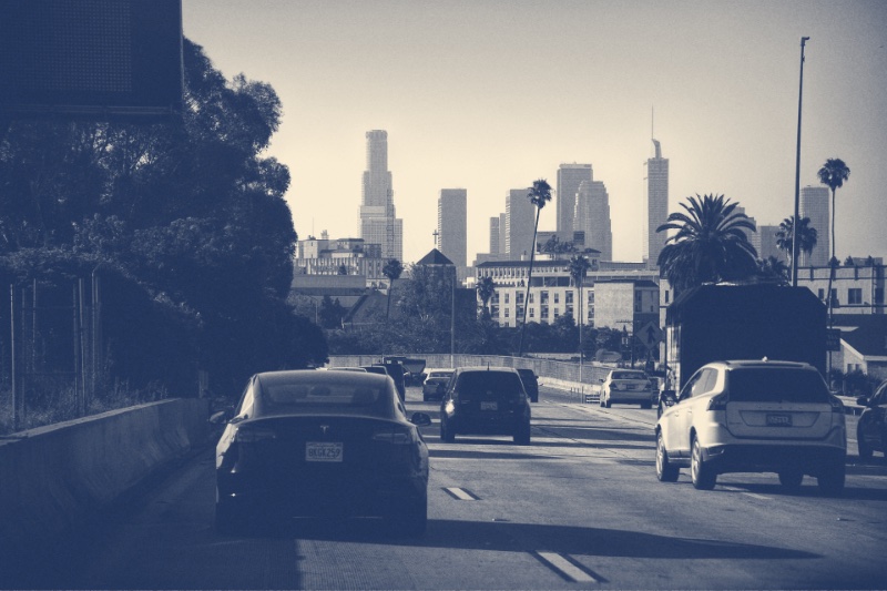 Cars on a Los Angeles freeway heading toward the downtown towers, an electric sedan in the near lane, in a slate and bone duotone