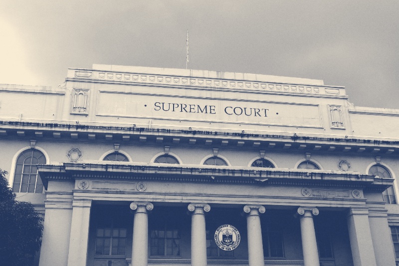 The facade of the Supreme Court of the Philippines on Padre Faura Street in Manila, the words Supreme Court carved above a row of Ionic columns and the Court’s round seal between them, under a heavy grey sky, in a slate blue and bone duotone