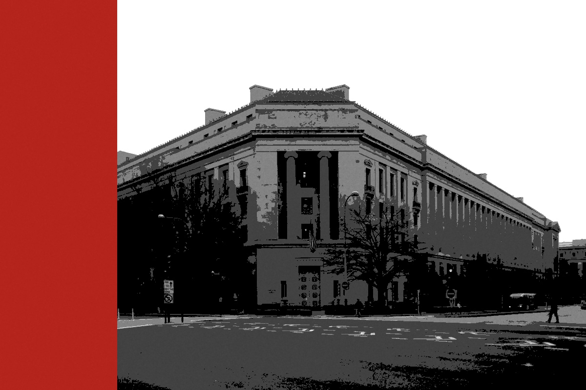The Robert F. Kennedy Department of Justice Building in Washington seen across an empty intersection, a flag over its corner entrance and bare trees along the pavement, in near black and white with a flat red bar down the left edge of the frame