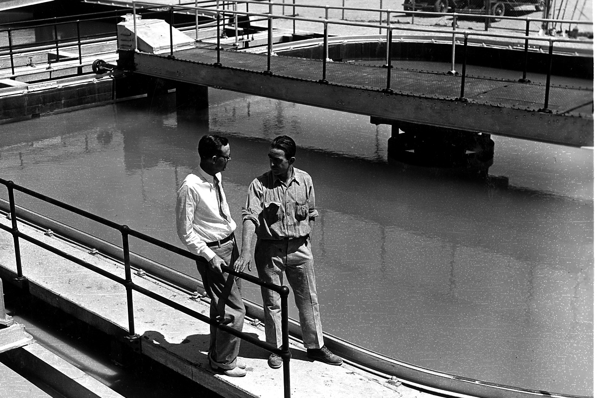 Two men in conversation on a walkway beside a circular water treatment basin, in plain black and white