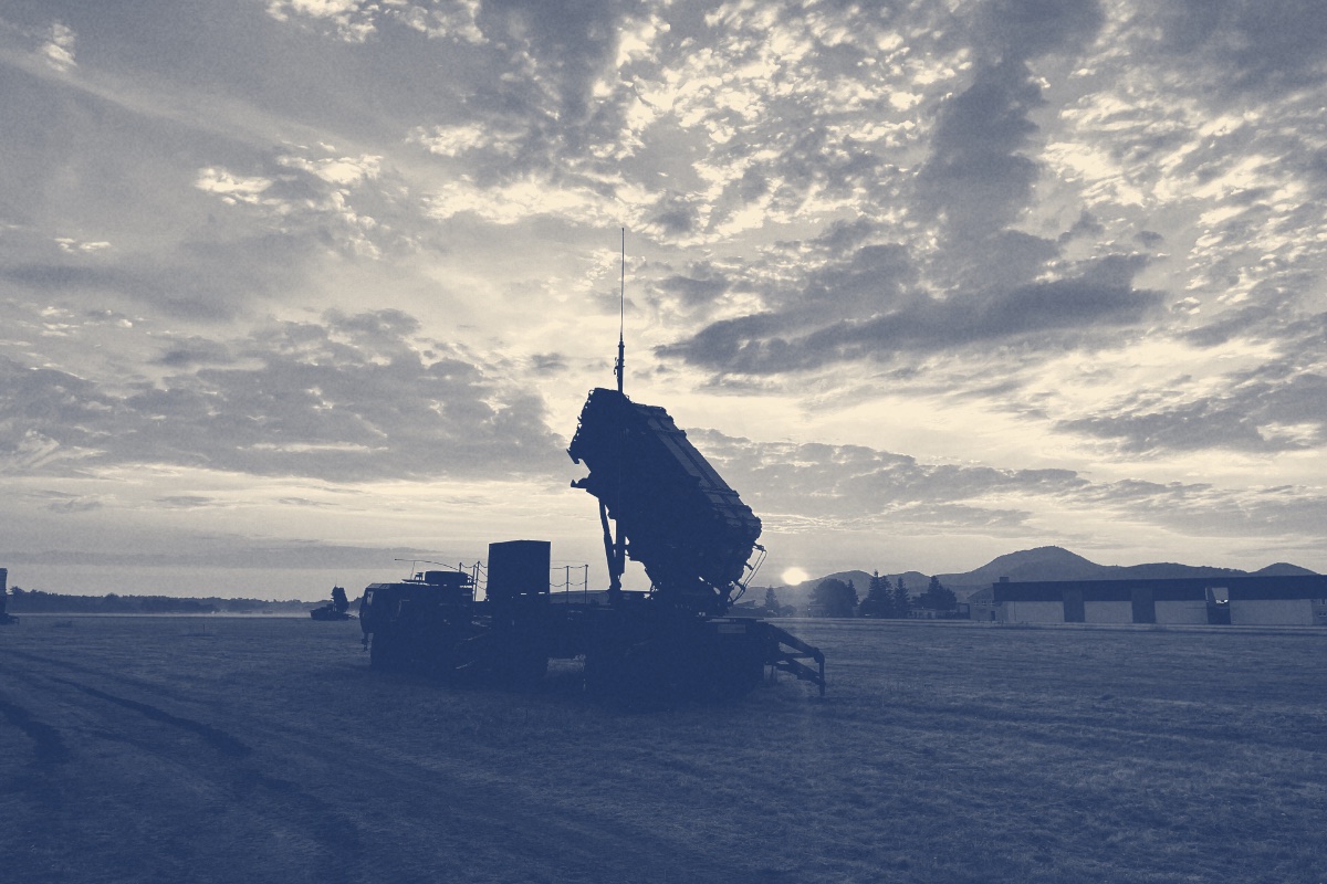 A Patriot missile launcher on its truck silhouetted against a sunrise over a flat grass field, low hills on the horizon under broken cloud, in a dark slate and bone duotone