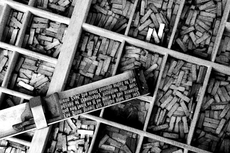 A compositor's case of metal type with a composing stick, in high contrast black and white with coarse grain