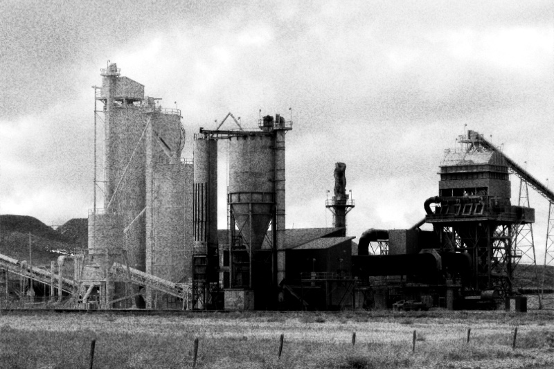 A lime plant in open country in Wyoming, silos, conveyors and a rotary kiln under an overcast sky behind a wire fence, in grainy high-contrast black and white