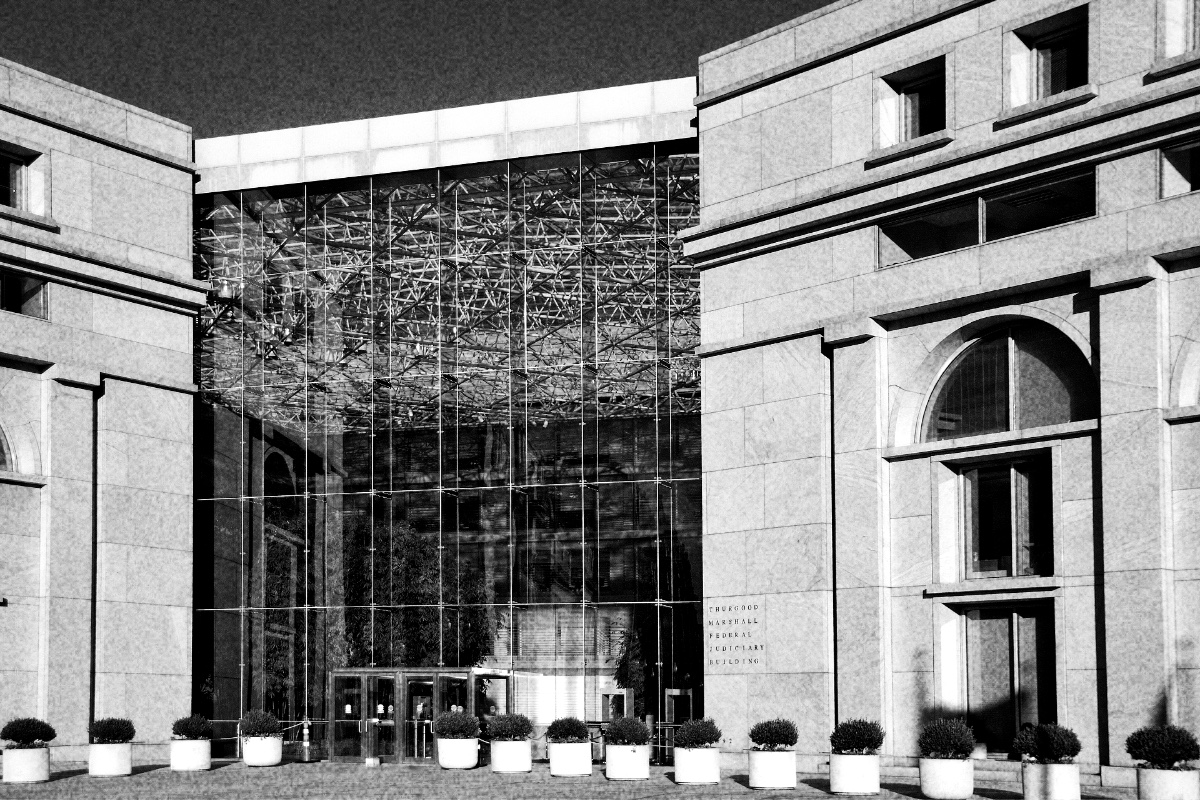 The glass atrium entrance of the Thurgood Marshall Federal Judiciary Building in Washington between two pale stone wings, with a row of potted shrubs along the pavement, in high-contrast grainy black and white