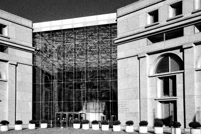 The glass atrium entrance of the Thurgood Marshall Federal Judiciary Building in Washington between two pale stone wings, with a row of potted shrubs along the pavement, in high-contrast grainy black and white