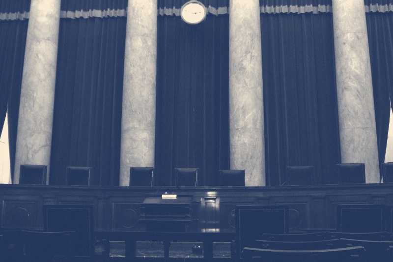 The empty courtroom of the U.S. Supreme Court, the justices' high-backed chairs behind the long bench, marble columns and heavy drapes behind them and a clock hanging above, in a slate and bone duotone