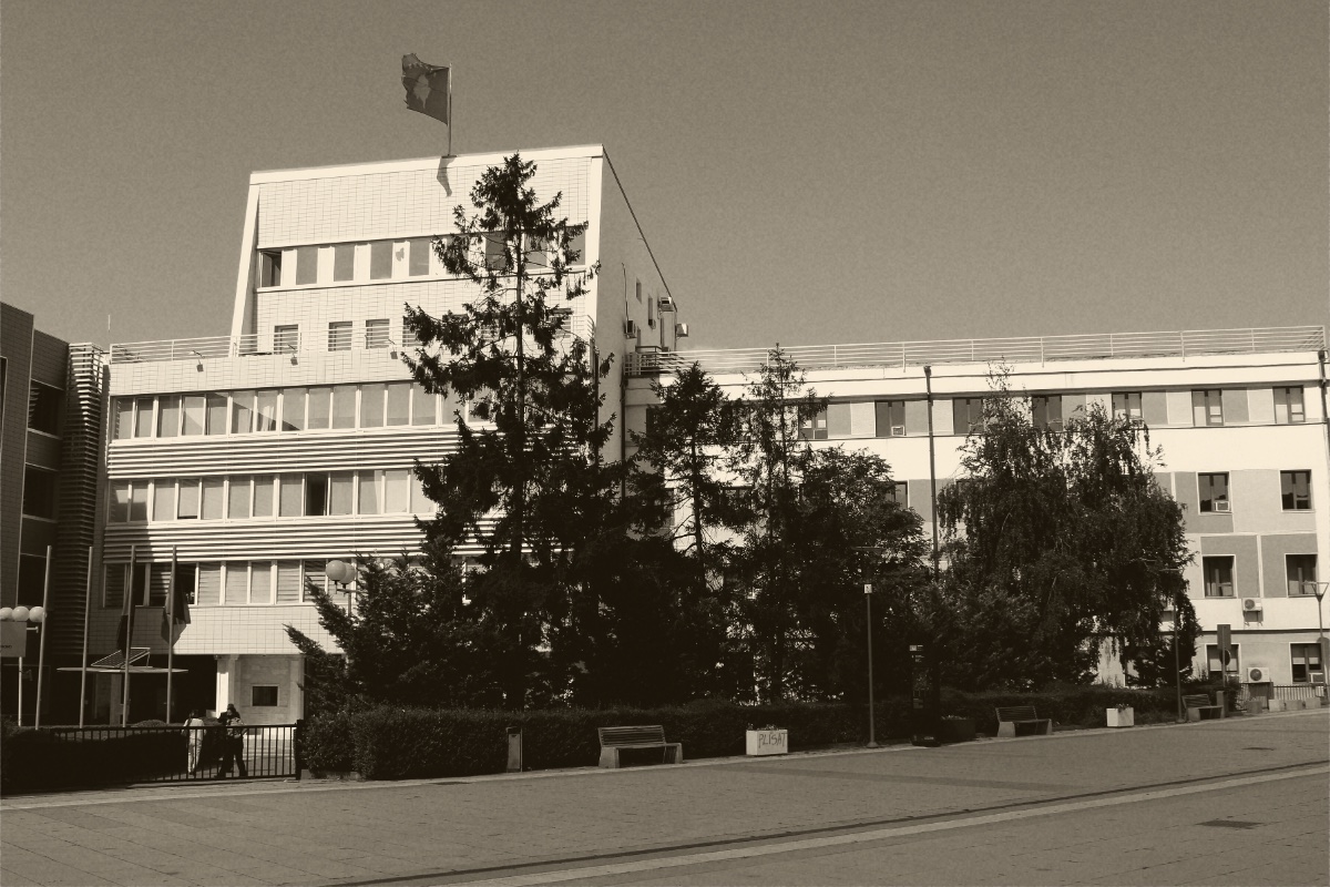 The Assembly of the Republic of Kosovo in Pristina, a pale multi-storey building with a flag on its roof behind a line of trees, in an ink and bone duotone