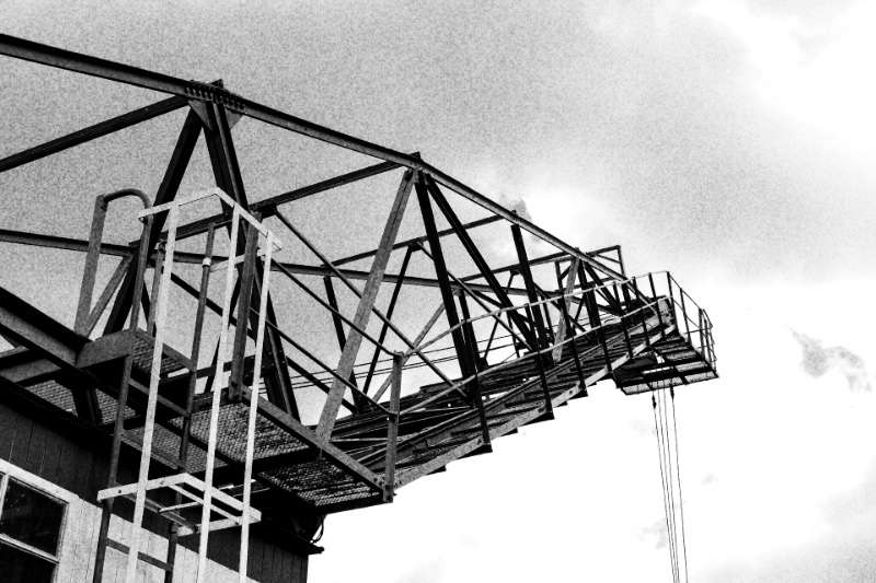 A steel harbour crane seen from below against the sky, in high contrast black and white with coarse grain