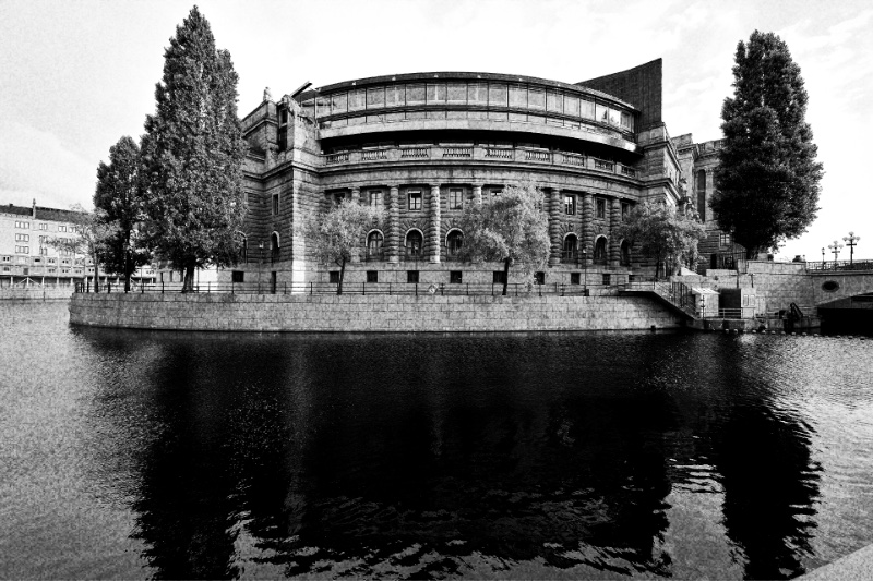 The Riksdag building in Stockholm seen across the dark water of the Stallkanalen, a curved stone and glass wing behind a granite quay, with tall poplars at either side, in high-contrast grainy black and white