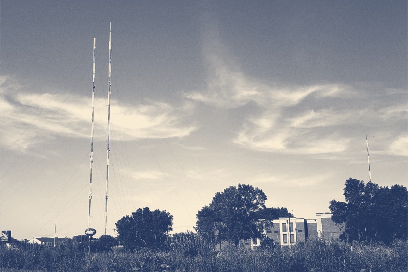 Two tall red and white broadcast masts beside a low brick building and a line of trees in Shoreview, Minnesota, under a wide sky, in a slate and bone duotone