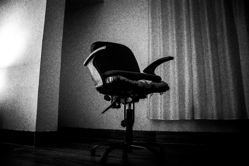 An empty, worn office chair with torn upholstery standing alone by a curtained window, in high contrast black and white with coarse grain