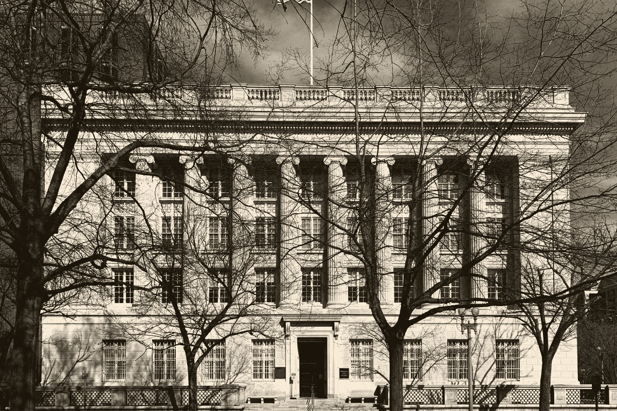 The Treasury Annex, now the Freedman’s Bank Building, in Washington, a pale stone facade of tall columns seen through bare winter trees, a flag on the roof, in a dark ink and bone duotone