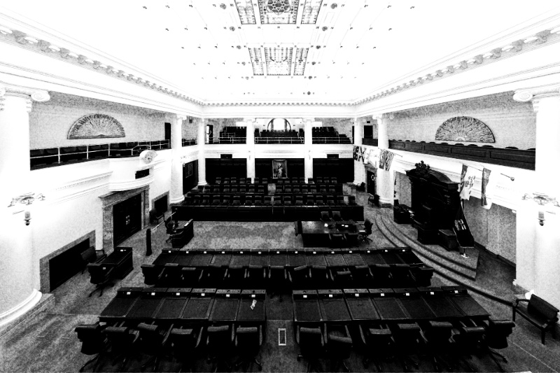 The empty chamber of the Legislative Assembly of Alberta seen from the public gallery, rows of desks and leather chairs facing the Speaker’s carved chair under a coffered ceiling with stained-glass panels, in grainy high-contrast black and white