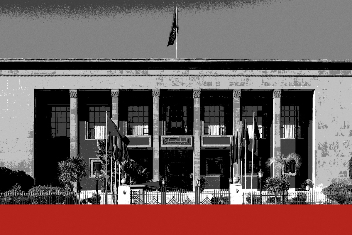 The parliament building in Rabat, a flat-roofed stone facade with square pillars framing tall recessed windows, flags on poles behind a railing and a flag on the roof, with two pedestrians on the wet pavement in front, in near black and white with a flat red band along the foot of the frame