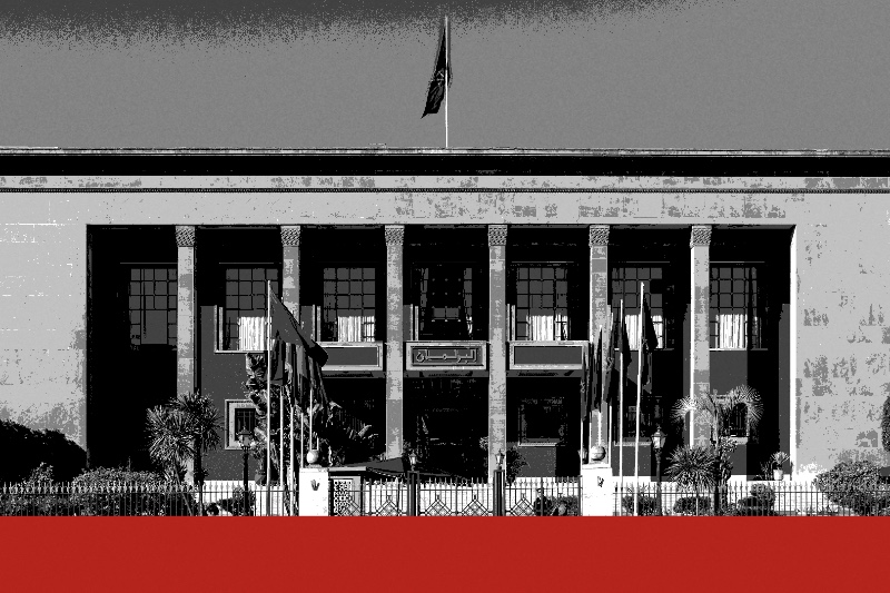 The parliament building in Rabat, a flat-roofed stone facade with square pillars framing tall recessed windows, flags on poles behind a railing and a flag on the roof, with two pedestrians on the wet pavement in front, in near black and white with a flat red band along the foot of the frame